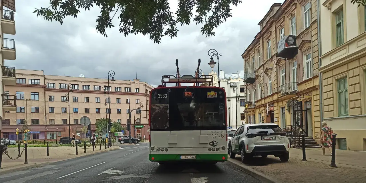 Trolejbus z numerem 3933 na tle kamienic i drzew. W tle widać fragmenty ulicy i innych pojazdów, a także słupy trakcyjne, które są częścią linii tramwajowej.
