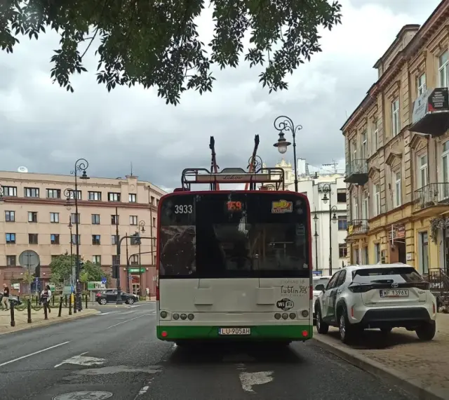 Trolejbus z numerem 3933 na tle kamienic i drzew. W tle widać fragmenty ulicy i innych pojazdów, a także słupy trakcyjne, które są częścią linii tramwajowej.