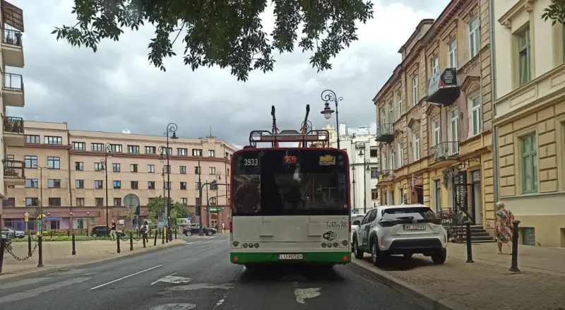 Trolejbus z numerem 3933 na tle kamienic i drzew. W tle widać fragmenty ulicy i innych pojazdów, a także słupy trakcyjne, które są częścią linii tramwajowej.